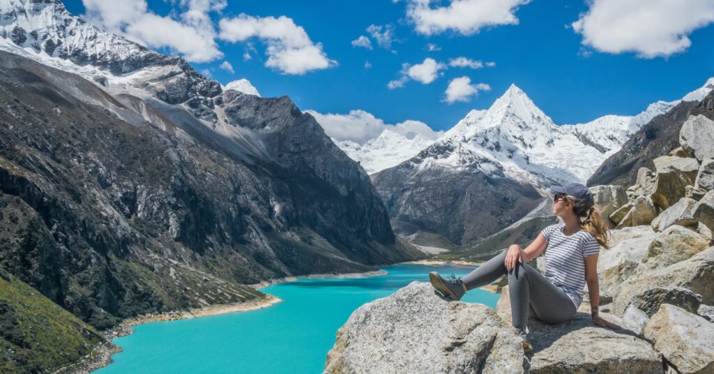 Female traveler sits on a rocky ledge gazing out over a turquoise mountain lake surrounded by snowy peaks, representing the stunning affordability and nature found in the cheapest countries to move in South America.