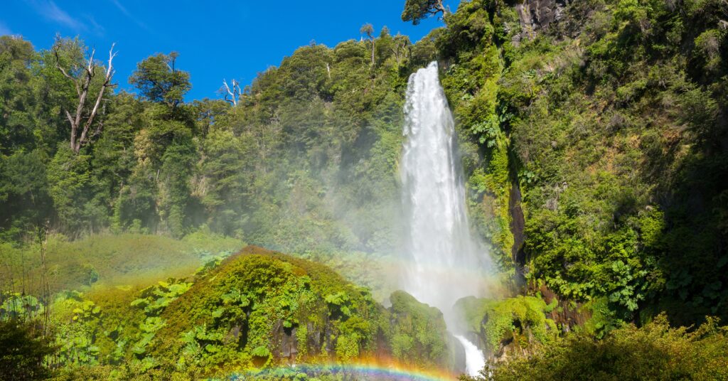 Majestic waterfall cascading down a lush green cliff surrounded by mist and a visible rainbow, showing the eco-diverse beauty awaiting those moving to Chile from the US.