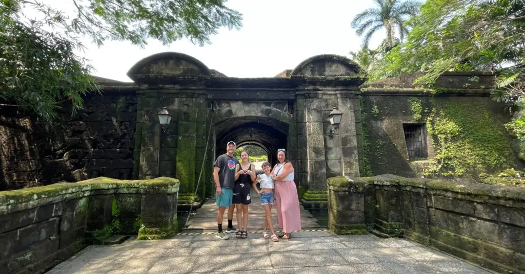 Smiling family of four stands together in front of a moss-covered stone fortress surrounded by tropical greenery, suggesting affordable adventure and quality of life in one of the cheapest countries to move as a family.