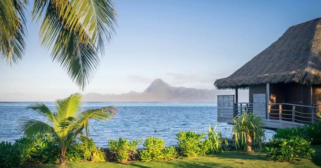 A tropical overwater bungalow with lush greenery and palm trees, overlooking a peaceful bay and distant mountains—an ideal scene representing affordable island living among the cheapest countries to move.