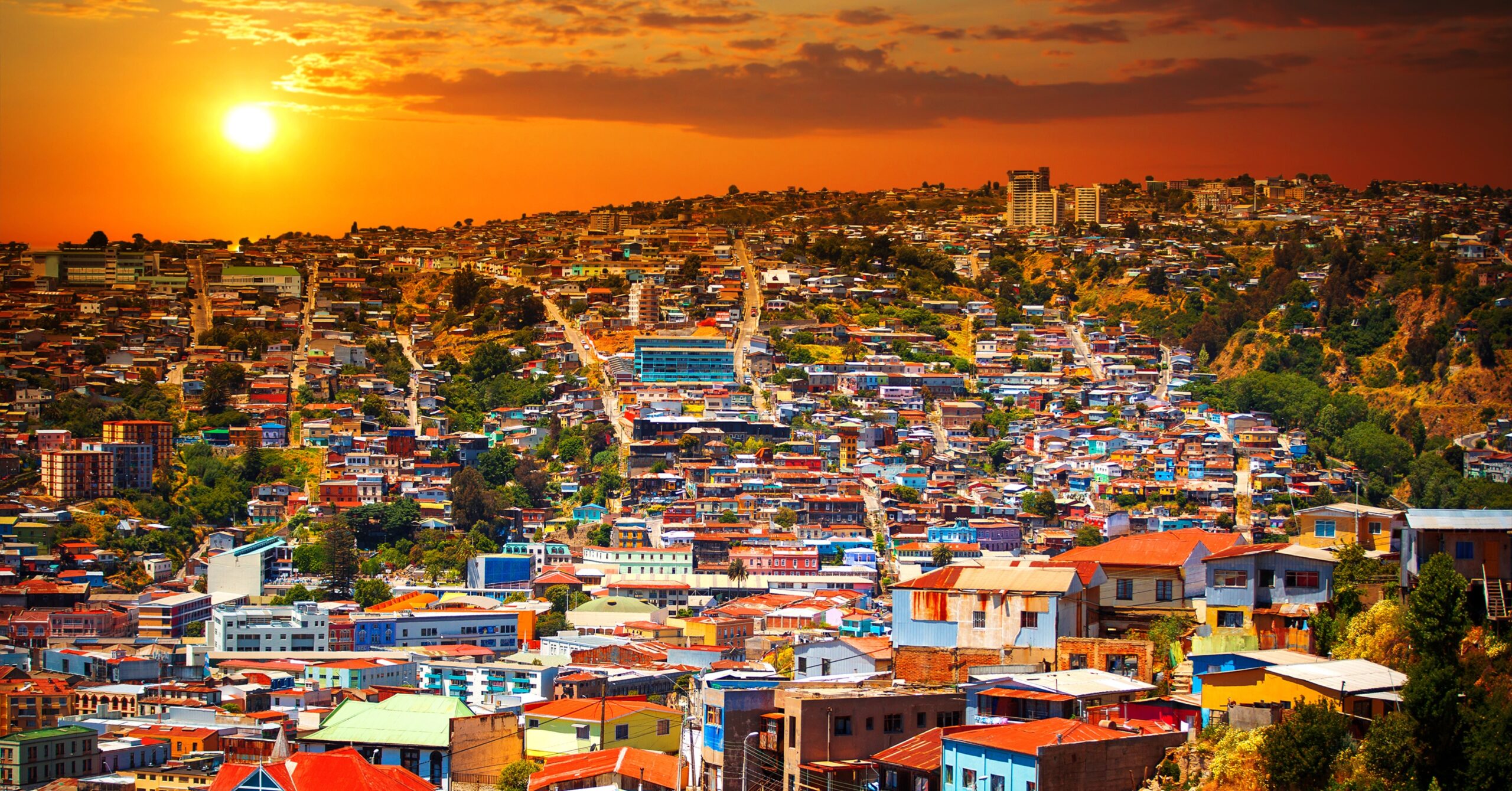Expansive view of Valparaíso's colorful hillside homes at sunset, with the sun glowing orange over the Pacific — a popular artistic and coastal destination for expats moving to Chile from the US.